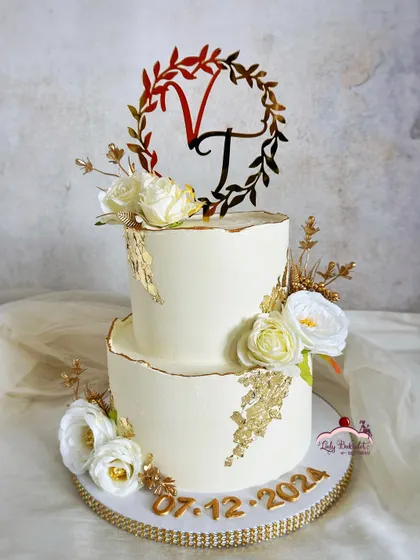 Gold accents and love-filled layers. An elegant two-tier engagement cake in ivory and gold, adorned with white roses and a custom monogram topper.
