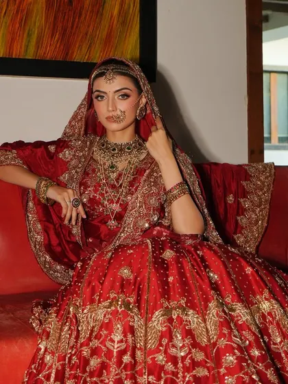 A regal portrait of a bride in a timeless red lehenga. Seated on a vintage sofa, the styling evokes a sense of heritage and poise, highlighting the rich embroidery and traditional jewelry.