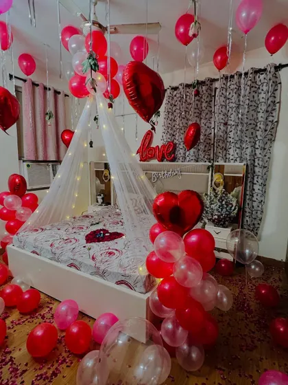 I design romantic room surprises for anniversaries and special moments. This setup features a fairy light canopy over the bed, with red and white balloons, heart-shaped foil balloons, and rose petals to create a truly memorable experience.