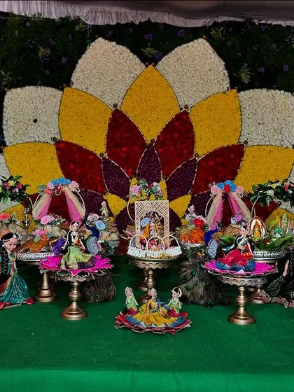 A vibrant and colorful gift platter setup for a half-saree function. The display features large, traditionally dressed dolls and a backdrop with a floral lotus design.