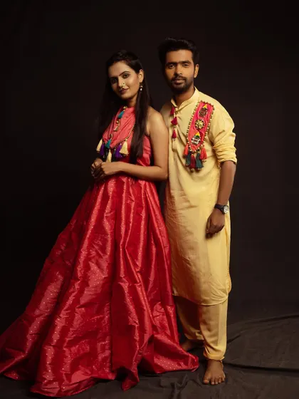 A full-length portrait showing off the complete festive look. The woman's flowing red gown and the man's yellow kurta create a striking contrast against the dark background.