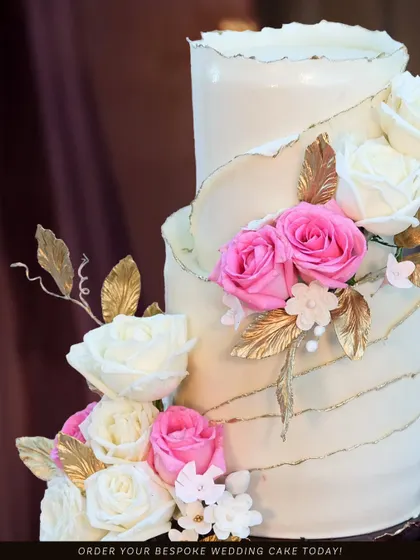 A detailed shot of an elegant wedding cake, adorned with fresh pink and white roses and delicate golden leaves. The torn-edge effect on the top tier adds a touch of modern texture.