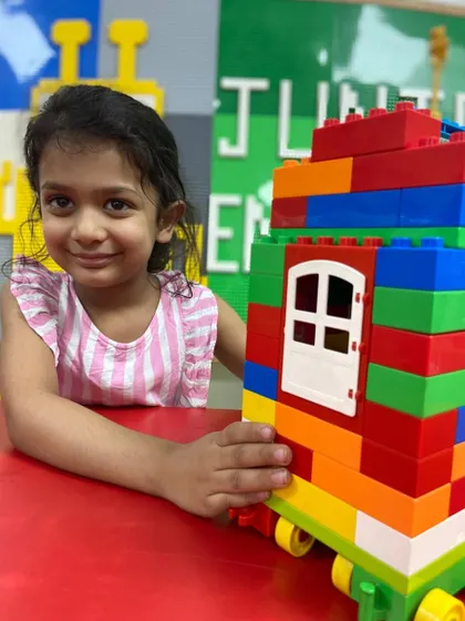A little girl smiles with her multi-colored house built from large DUPLO-style blocks, showcasing her early construction skills.