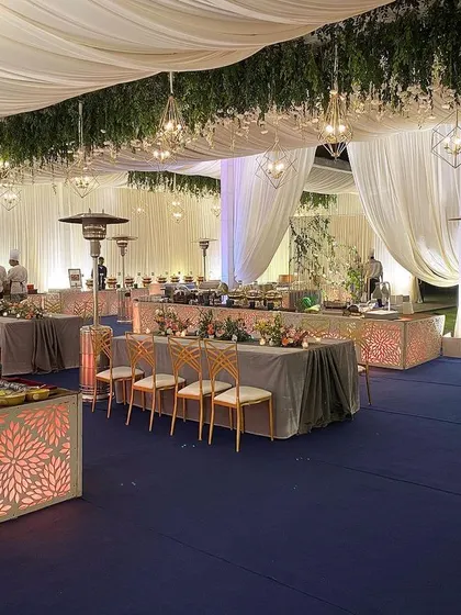 A wide view of an elegant reception buffet area. The space is defined by a deep blue carpet and clean white drapes, with greenery and geometric chandeliers hanging from the ceiling to add a touch of modern sophistication.
