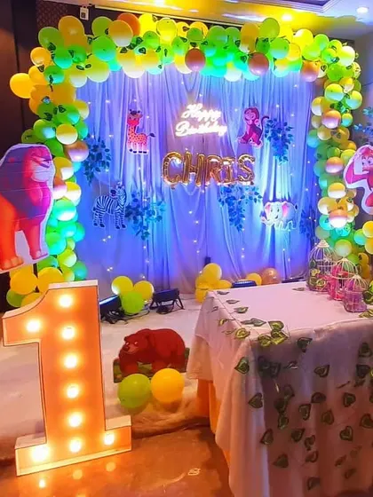 A vibrant jungle-themed first birthday setup. The decor includes a backdrop with a leafy curtain, animal cutouts like a lion and deer, and a light-up number '1'.