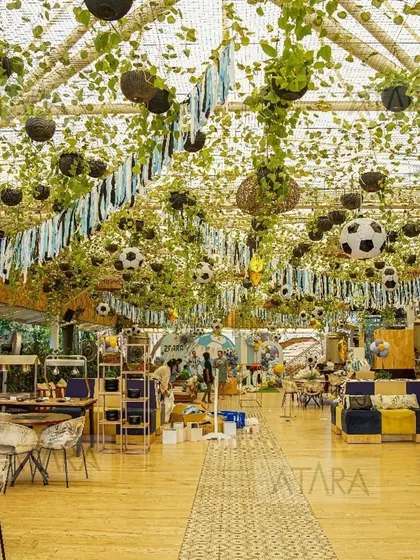 A wide shot of the indoor soccer-themed party, showing the full extent of the ceiling decor with hanging plants and soccer balls. The wooden floors and furniture give the space a rustic yet sporty feel.