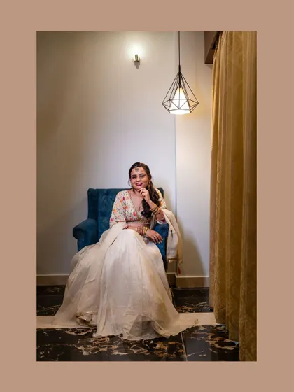 A portrait of the bride sitting in a blue armchair, her white lehenga spread out beautifully around her.