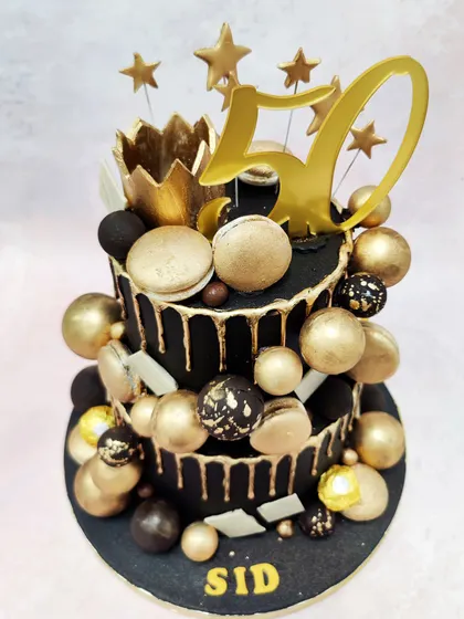 We mark a golden jubilee with this spectacular black and gold 50th birthday cake. Two tiers of black fondant with gold drips are lavishly decorated with a gold crown, stars, macarons, and a cascade of gold and black spheres for a truly grand celebration.