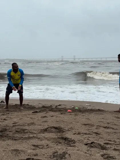 Players listen intently to coaching instructions during a beach fielding drill, with the challenging waves and sand providing a unique training backdrop.
