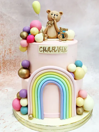 We combine several whimsical themes in this two-tiered rainbow teddy bear cake. A sweet teddy bear holding a balloon sits atop a pink cake adorned with a pastel rainbow and a cascade of colorful spheres, creating a joyful and imaginative design.