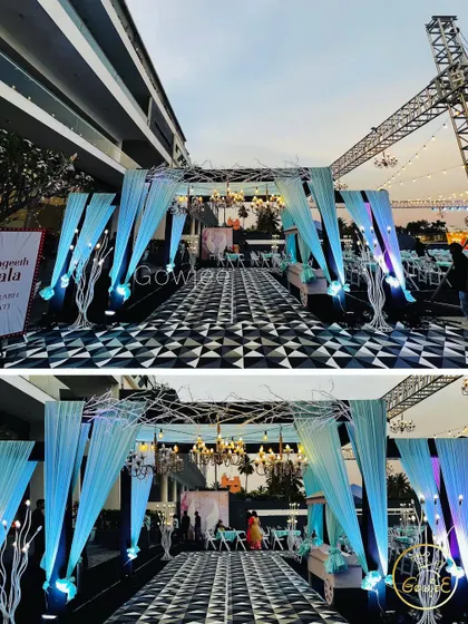 A modern Sangeet entrance with a checkered floor, light blue drapes, and artistic white tree props.