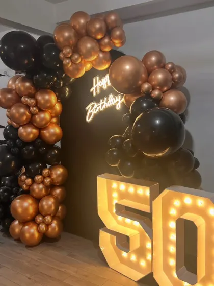 This view highlights the full curve of the balloon arch as it frames the backdrop. The mix of different sized balloons in metallic bronze and classic black adds texture and a modern, sophisticated feel.