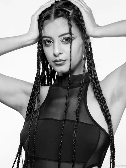 A high-contrast black and white portrait of a model with long braids and graphic eyeliner. The white background makes the subject pop, creating a striking editorial image.