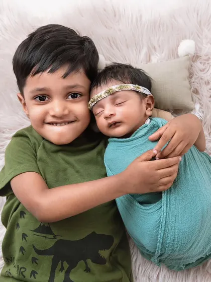 A proud big brother smiles for the camera while hugging his swaddled newborn sibling. Capturing the personalities of both children is key to a successful sibling photoshoot.