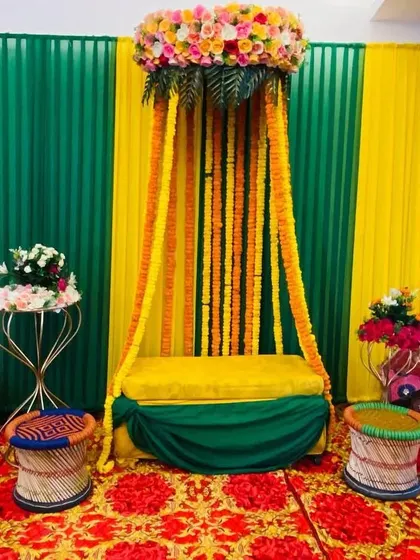 A traditional Mehendi seating area with a canopy of marigold strings. The setup includes colorful 'mooda' stools and a backdrop of green and yellow drapes.