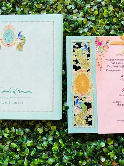A beautiful pastel wedding invitation featuring a traditional peacock and Ganesha motif. The card has a laser-cut panel with gold foil accents and a ribbon tie, blending classic Indian design elements with a soft, modern color scheme.