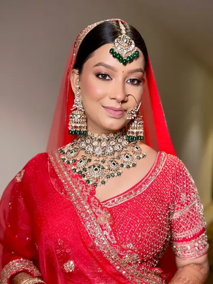 A close-up of a bride in a classic red lehenga. This shot perfectly showcases the heavy Kundan and green bead jewellery set, including the oversized jhumkas and matching maang tikka.