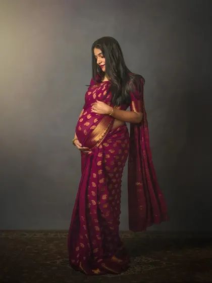 A full-length studio portrait of an expecting mother in a gorgeous magenta saree. The simple, dark backdrop makes the rich color and gold details of the fabric stand out, emphasizing her graceful silhouette.