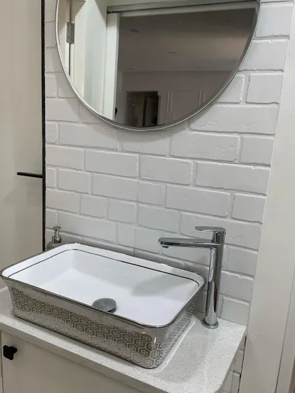A close-up of a modern washbasin area in a common space. The design features a stylish rectangular vessel sink with a geometric pattern, set against a white subway tile wall and complemented by a simple round mirror.