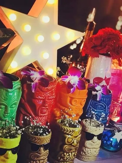 A collection of colorful tiki-style mugs and other retro decor. The glowing star marquee and vibrant flowers add to the fun, nostalgic feel of this 80s-themed cocktail bar.
