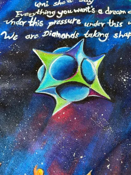 A close-up of the hand-lettered lyrics and the geometric star shape from the "Adventure of a Lifetime" artwork. The white lettering on the dark, starry background creates a powerful visual contrast.