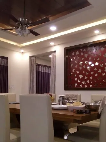 An elegant dining space designed for family meals, featuring a large wooden table with upholstered chairs. The room is enhanced by a wooden false ceiling panel, cove lighting, and a distinctive red decorative wall panel with a metallic leaf motif.