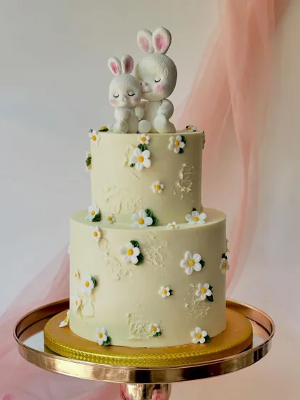 A two-tiered cake with two adorable bunnies cuddling on top. The cake is decorated with small, delicate daisy flowers for a soft and gentle look.