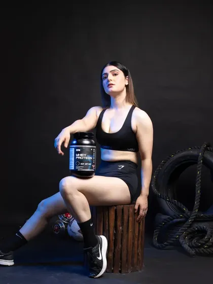 A female model in athletic wear holding a tub of whey protein, seated in a gym setting with tires and ropes to create a rugged, authentic fitness vibe.