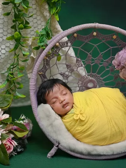 A newborn wrapped in yellow, sleeping in a purple dreamcatcher swing. The green background makes the colors stand out.