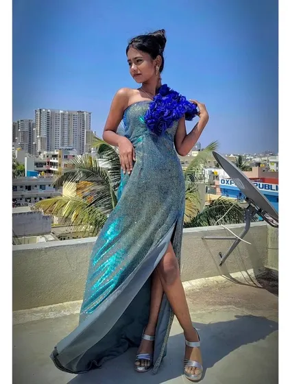 A model posing on a rooftop in a shimmering high-slit gown with a blue floral shoulder piece.