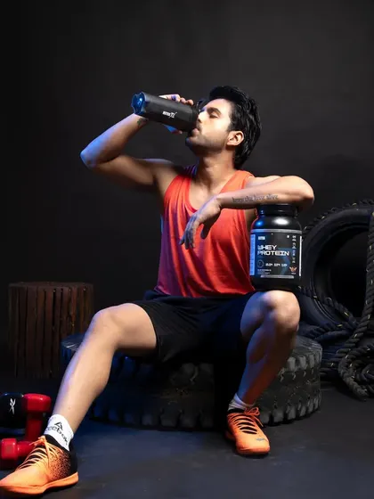 A male model relaxing post-workout, drinking from a shaker bottle with a tub of whey protein by his side. This image tells a story of product use and satisfaction.