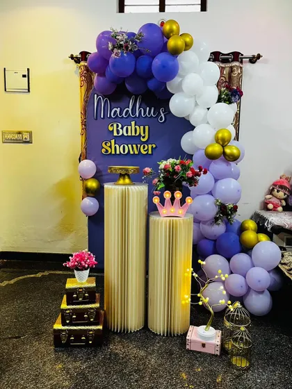 An intimate and stylish baby shower corner setup. This decoration features a purple backdrop, a white and purple balloon garland with gold accents, and elegant fluted plinths for placing cakes or gifts.