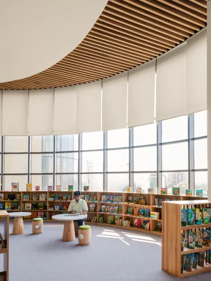 A quiet reading corner in the Vedanya School library, flooded with natural light from floor-to-ceiling windows. The curved bookshelves follow the building's organic form, creating a peaceful and inspiring atmosphere.