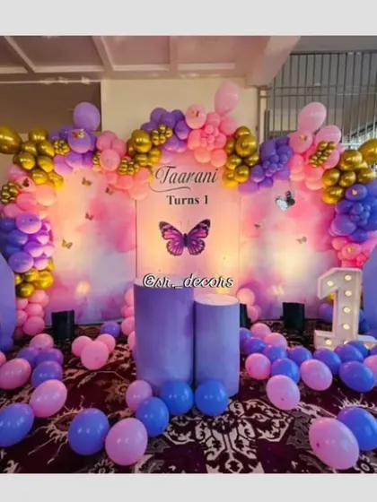 A grand butterfly-themed stage for a first birthday, using a colorful backdrop, large decorative butterfly wings, and a multi-toned balloon arch in purple, pink, and gold.