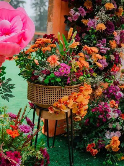 A beautiful, lush floral arrangement in a cane planter, featuring a mix of orange, pink, and purple blooms. The giant pink paper flower in the background adds a touch of whimsical scale.