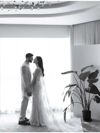 A wide, minimalist black and white shot of a couple about to kiss, their figures framed by a large window and a simple houseplant. A moment of quiet intimacy.