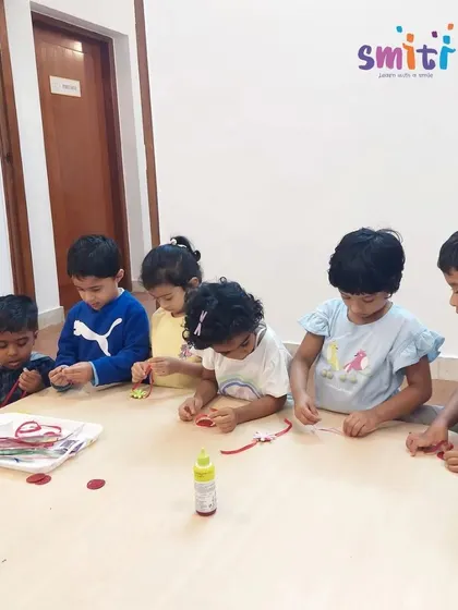 Children sit at a table, focused on gluing decorations onto their handmade Rakhis. This activity requires concentration and develops fine motor skills.