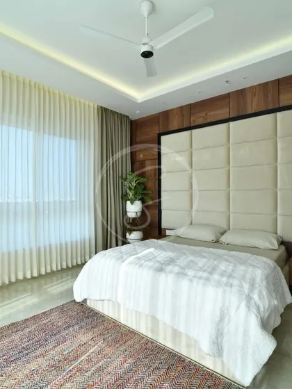 A minimalist bedroom with a rustic theme, featuring a grand, cream-colored upholstered headboard against a dark wood paneled wall. The design is clean and bright, with large windows and sheer curtains.