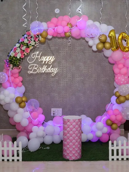 A trendy circular backdrop for a 10th birthday party. The ring is made of pink and white balloons with floral accents and features a "Happy Birthday" neon sign, creating a perfect photo booth area.