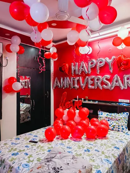 A simple anniversary room decoration with red and white balloons. The wall features 'Happy Anniversary' foil letters, perfect for a home celebration.