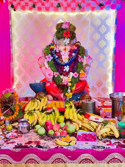 A traditional Ganpati decoration with a focus on the offerings. The idol is adorned with a large flower garland, and the stage is filled with fruits and prasad.