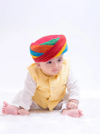 This baby looks absolutely adorable in a traditional Rajasthani outfit, featuring a colorful turban and a yellow vest. The simple white background makes the vibrant colors pop.