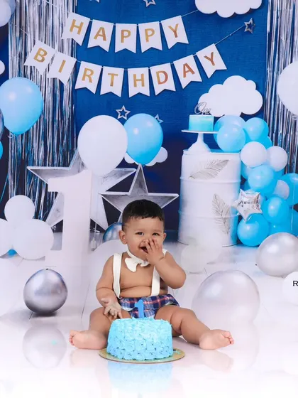 A classic cake smash pose with the birthday boy enjoying his first taste of cake. The blue and silver balloon backdrop creates a celebratory and stylish setting.