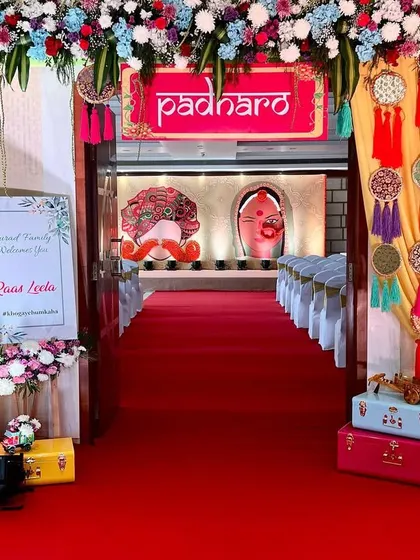 A vibrant and festive entrance for a 'Raas Leela' themed event. A bright red carpet leads guests through a floral archway towards a stage with traditional motifs.