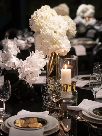 A luxurious table setting with a black reflective surface, gold-accented candle holders, and crisp white hydrangeas. The personalized acrylic menu adds a modern, bespoke touch.