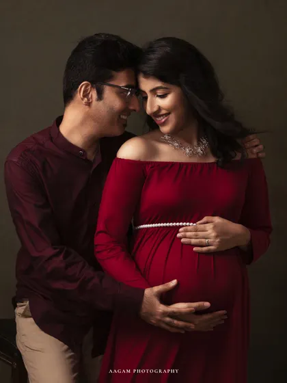 An expecting couple shares a loving gaze in this intimate fine art portrait. The deep red of her dress contrasts beautifully with the muted green backdrop, highlighting their connection.