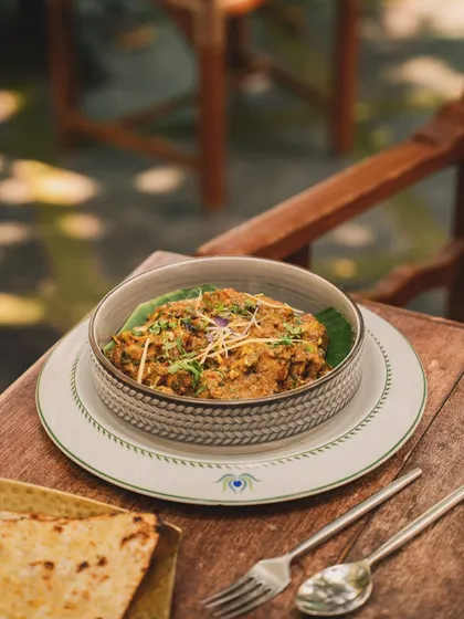 A rustic bowl of a savory dish, served with flatbread on a wooden chair. The outdoor, dappled sunlight gives this photo a warm, relaxed, and al fresco dining feel.