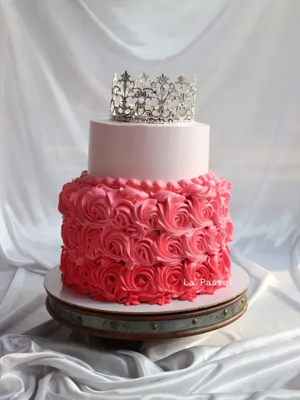 A cake fit for a princess! This two-tier design features a smooth pink top tier and a bottom tier covered in beautiful pink ombre rosettes, all topped with a silver crown.