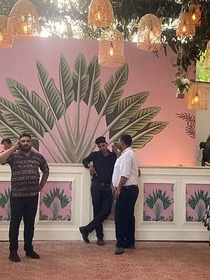 A custom-designed photo wall for a tropical-themed Mehendi. The hand-painted pink backdrop with large banana leaves adds a fun, artistic, and highly photogenic element to the event decor.
