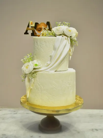 An elegant two-tier engagement cake with a subtle, embossed leaf pattern on the buttercream. The design is elevated with a flowing white fondant drape and fresh white flowers, complemented by a custom gold initial topper.